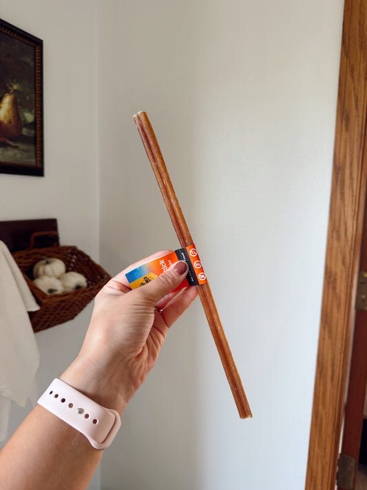 Hand holding a dog bully stick with a colorful label against a neutral background.