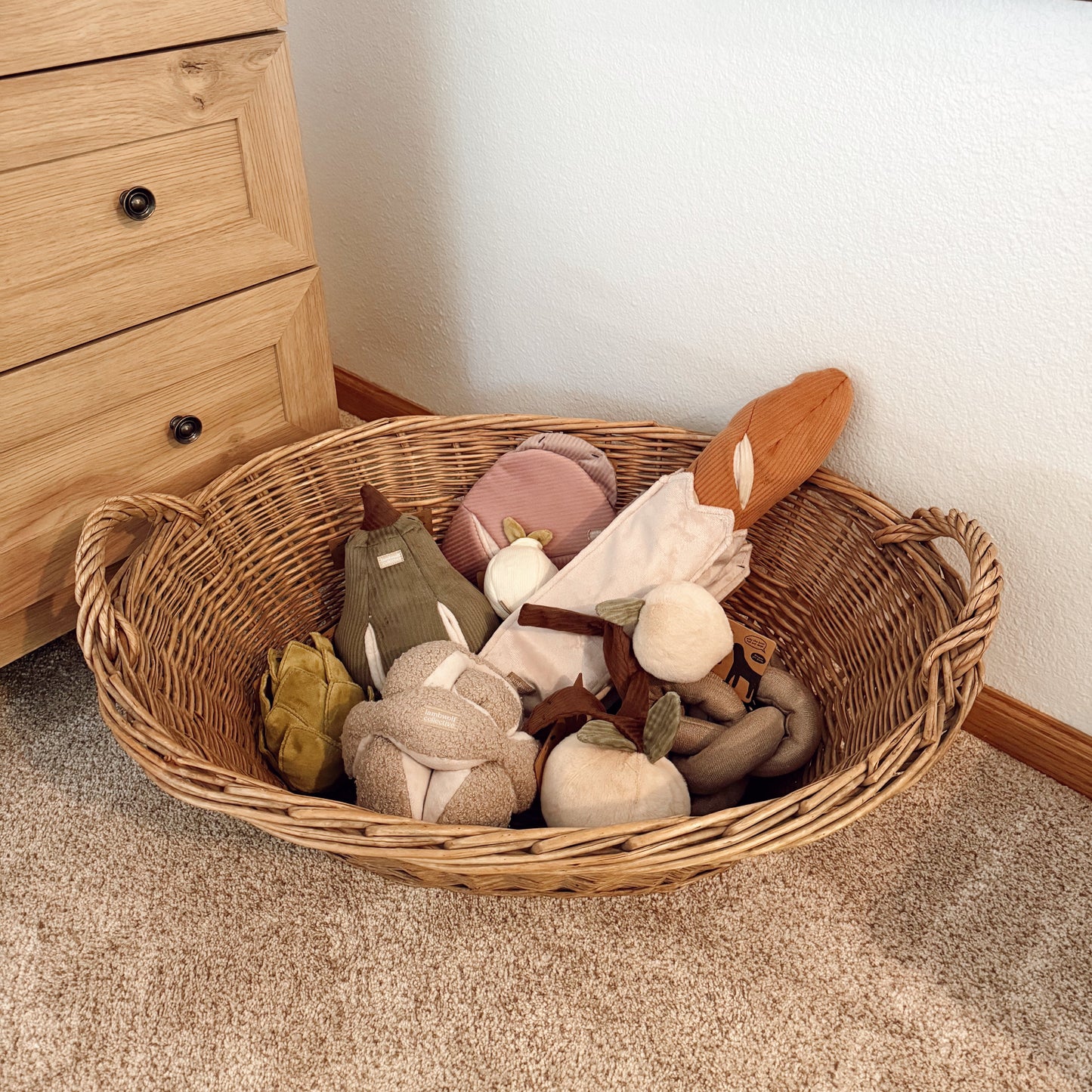 Wicker basket filled with soft toys on a carpeted floor next to a wooden dresser.