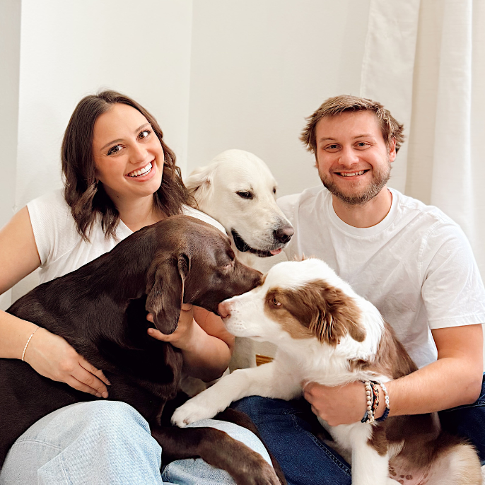 Two people sitting on the floor with three dogs in a bright room.