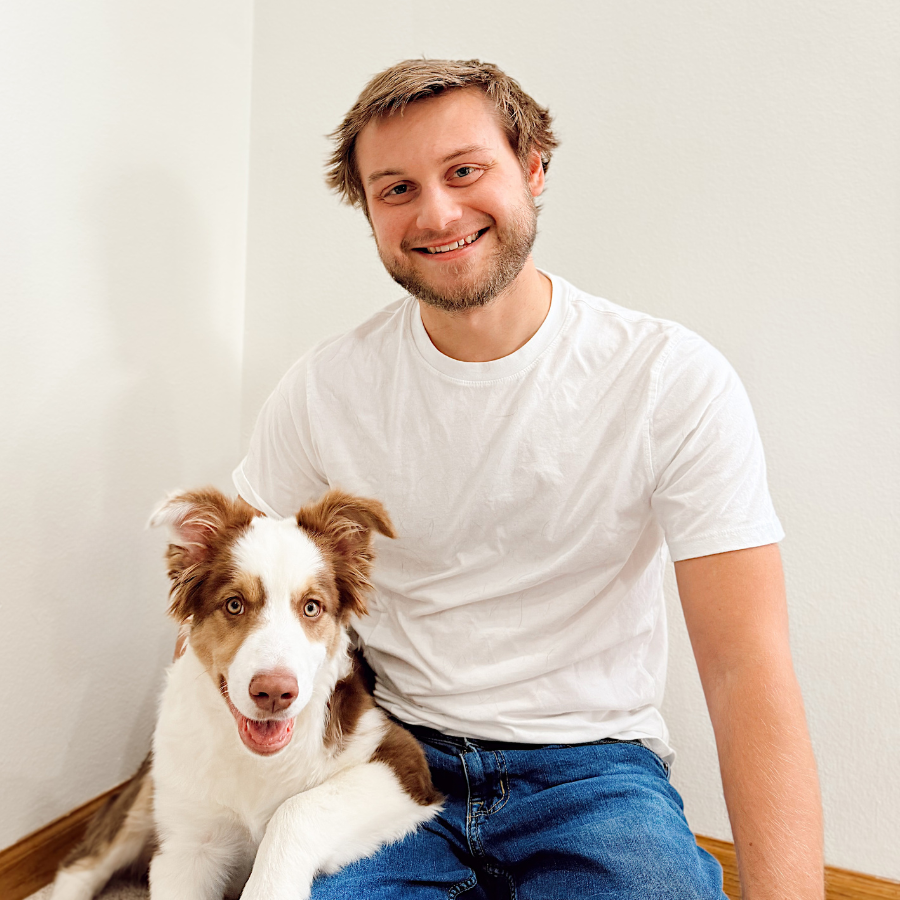 Man sitting with a dog on a carpeted floor against a white wall.