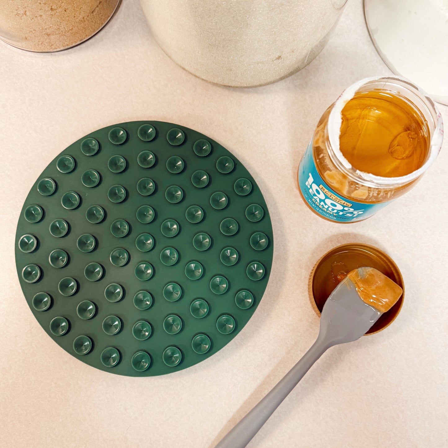 Green silicone dog lick mat upside down with a jar of peanut butter and a spatula on a kitchen countertop.