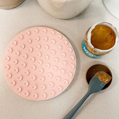 Pink dog lick mat on a kitchen counter upside down with peanut butter and a spatula.